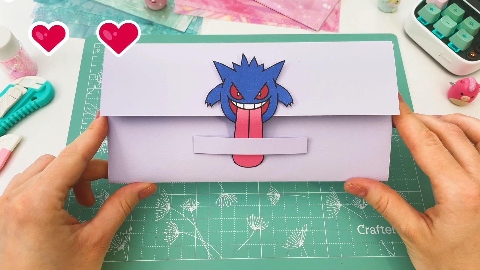 La billetera de papel terminada y cerrada, sostenida con las dos manos, con Gengar en la solapa y su lengua debajo de la banda