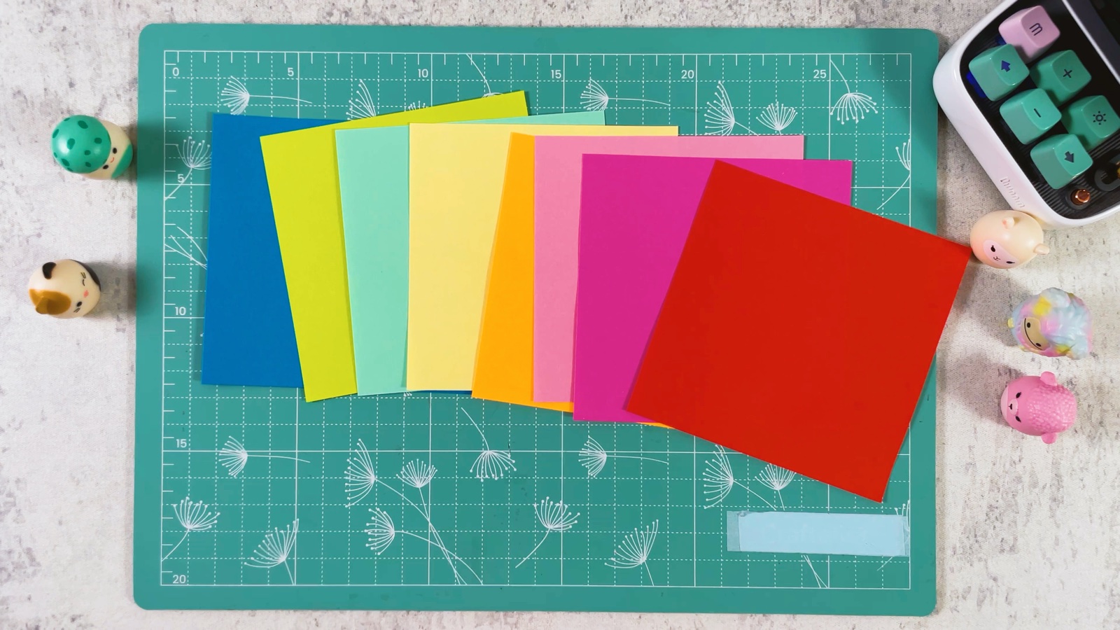 Eight square sheets of paper fanned out on a cutting mat in rainbow order, from blue through yellow and orange to pink and red