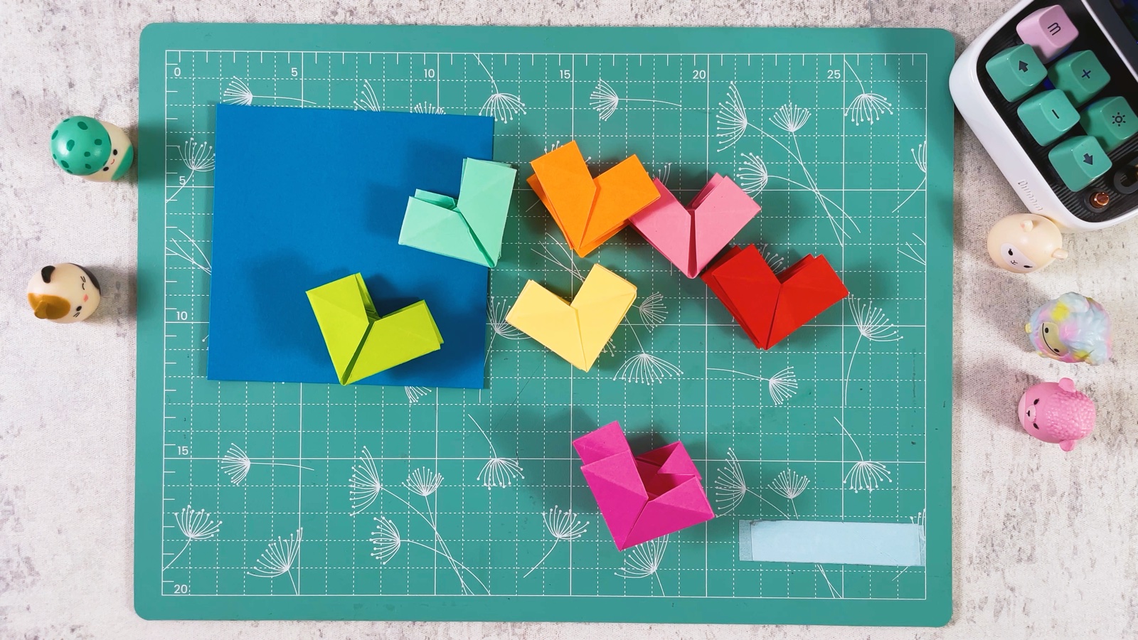 Folded paper hearts in green, mint, orange, yellow, pink and red scattered on the mat next to a blue square still waiting to be folded