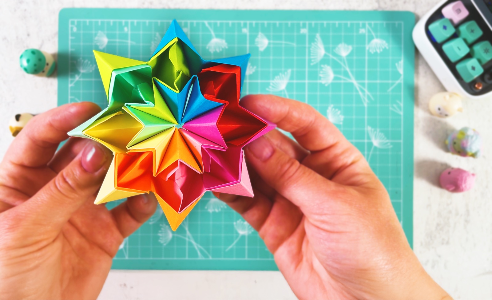 A finished rainbow origami infinity star held in two hands over a mint cutting mat