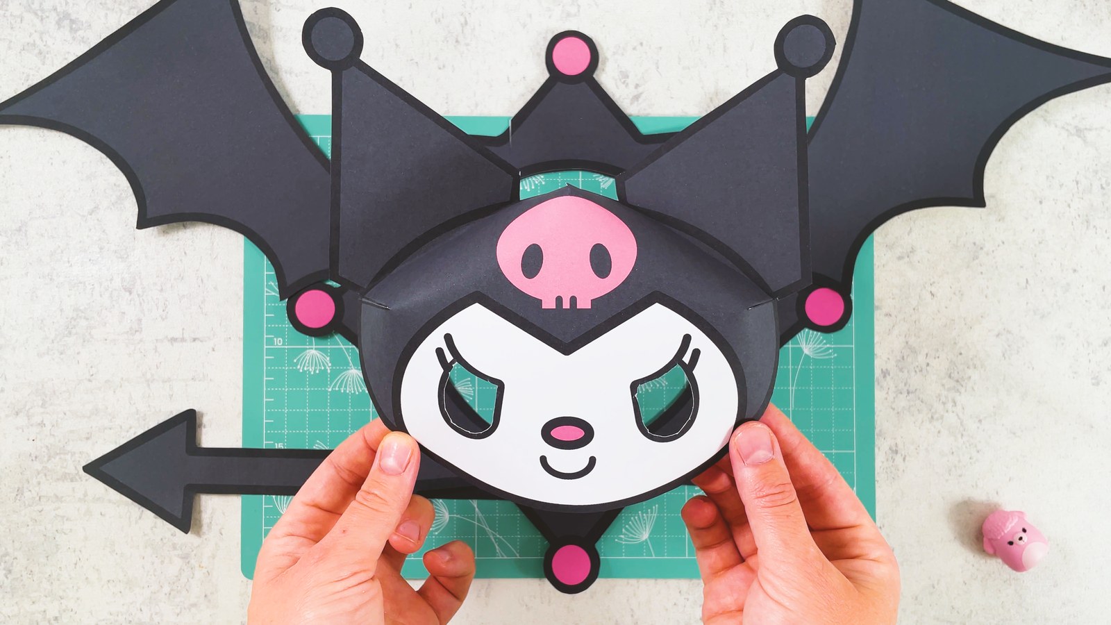 Das fertige Kuromi-Set aus Papier: die runde Maske mit rosa Totenkopf und spitzen Ohren in zwei Händen, dahinter der Zackenkragen, zwei Fledermausflügel und der Pfeilschwanz