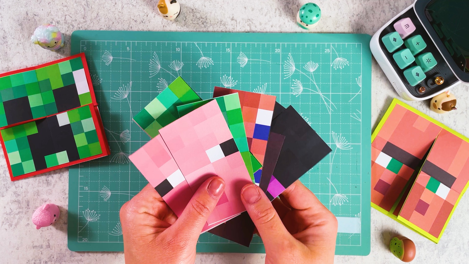 Printed Minecraft faces cut in half and fanned out in two hands: Pig, Creeper, Steve and Enderman