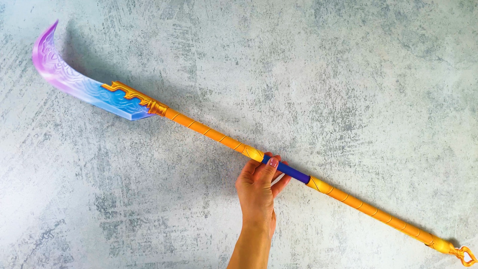 Das ganze fertige Gokdo aus Papier in einer Hand: eine gebogene rosa-blaue Klinge, ein langer goldener Stab mit lila Griff und eine goldene Spitze in Herzform