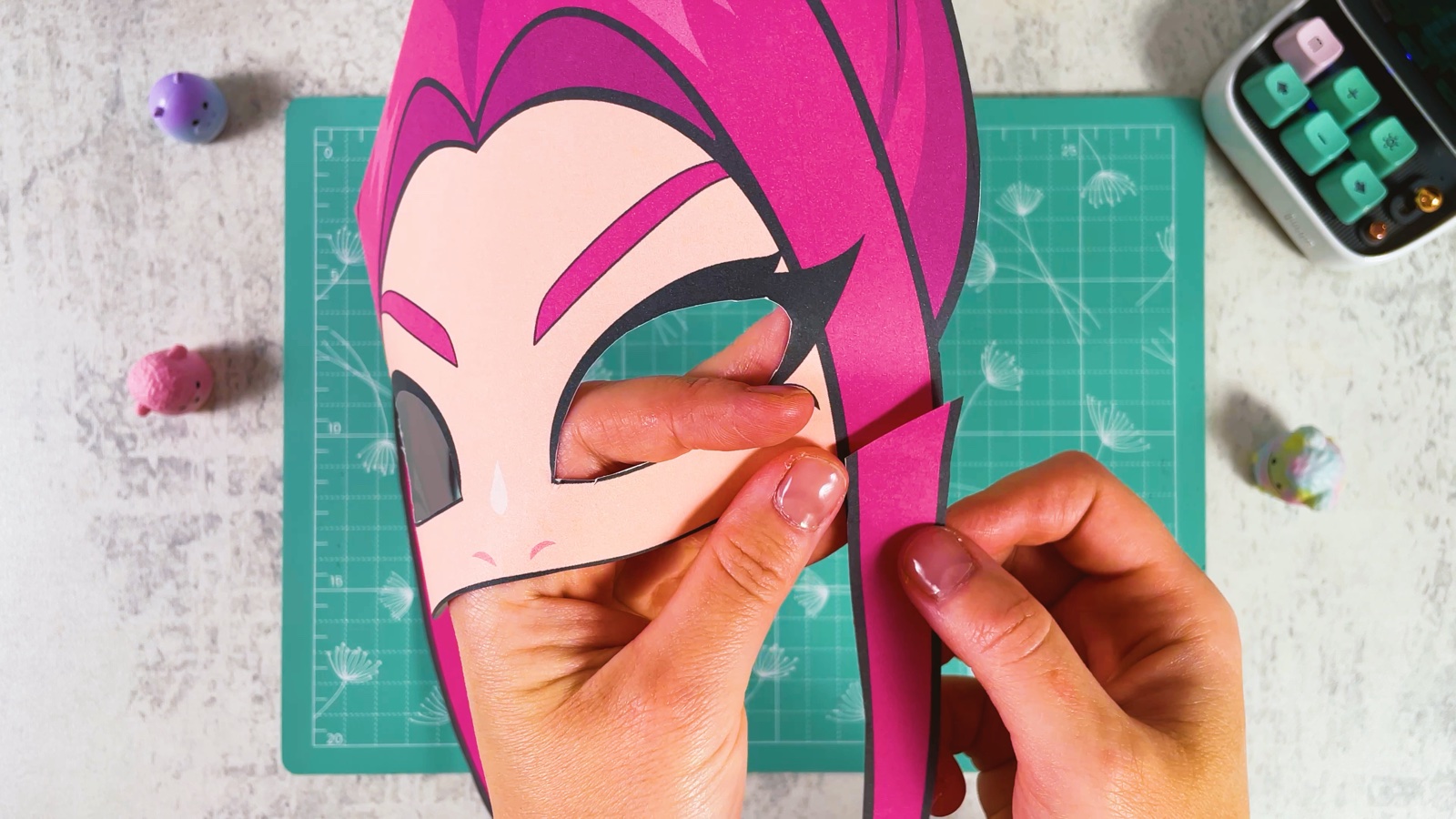 Pressing a long pink strand onto the side of the mask, next to Mira’s eye