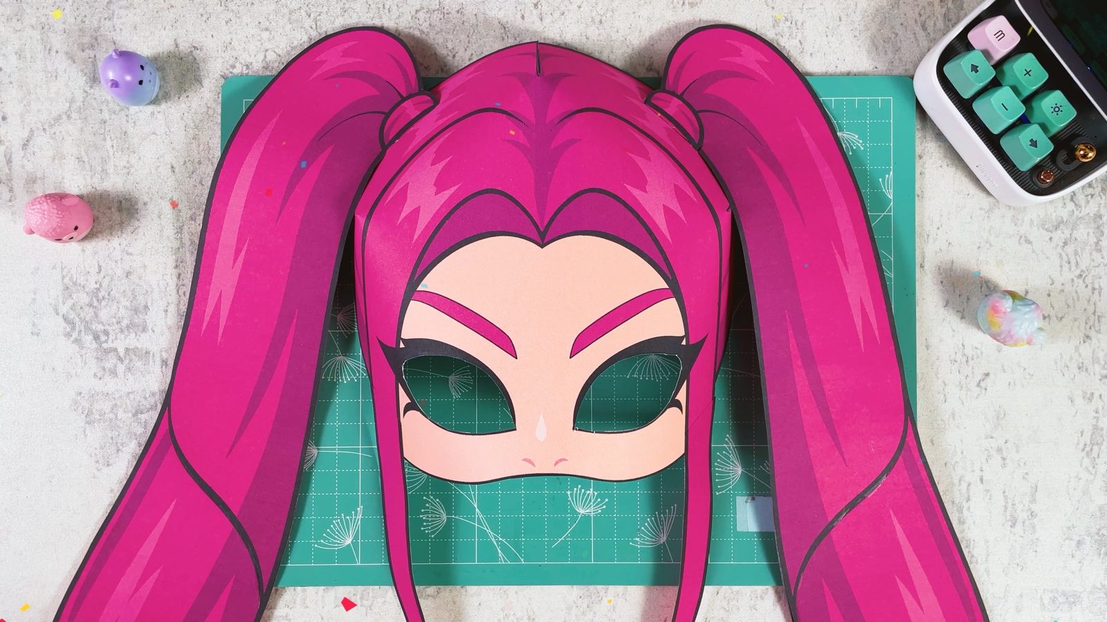 The finished paper Mira mask with long pink pigtails, lying on a mint cutting mat