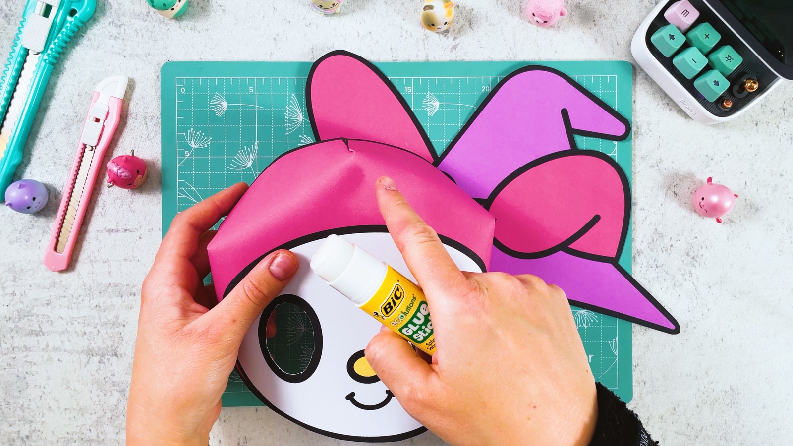 Mit einem Klebestift kommt Kleber oben auf die rosa Kapuze, dahinter wird der Hut hochgehalten