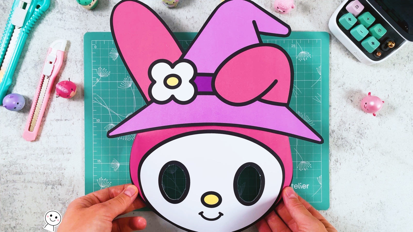 Die Maske mit dem aufgeklebten lila Hexenhut, das rosa Ohr steht nach oben
