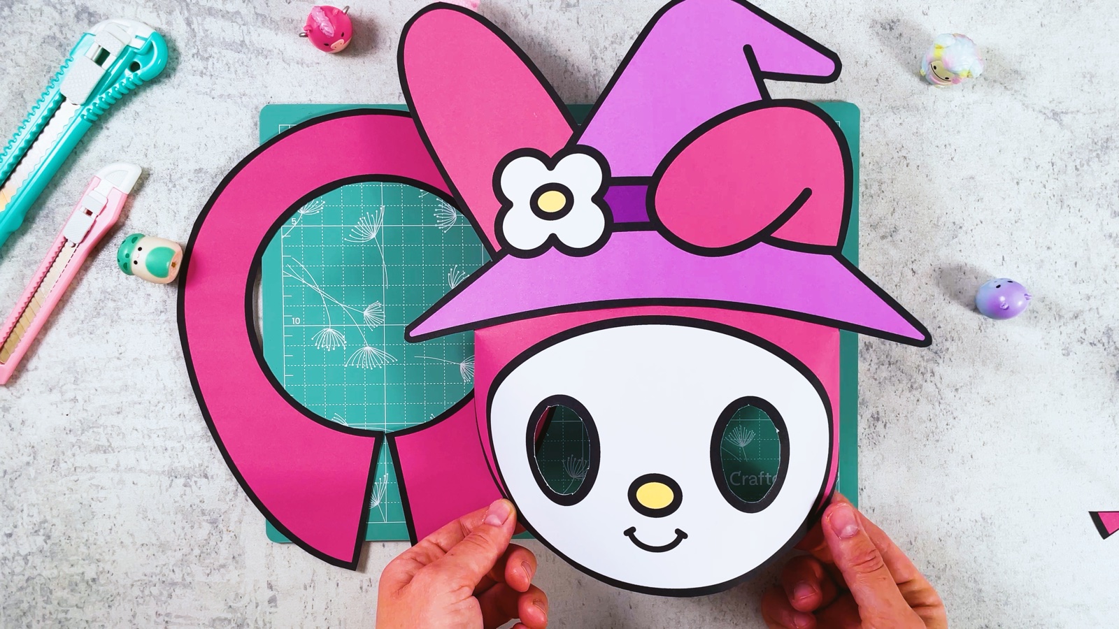 Die fertige My-Melody-Maske aus Papier wird hochgehalten, rund wie ein echter Kopf, mit lila Hexenhut, rosa Ohr und Schleife, dahinter der rosa Kragen