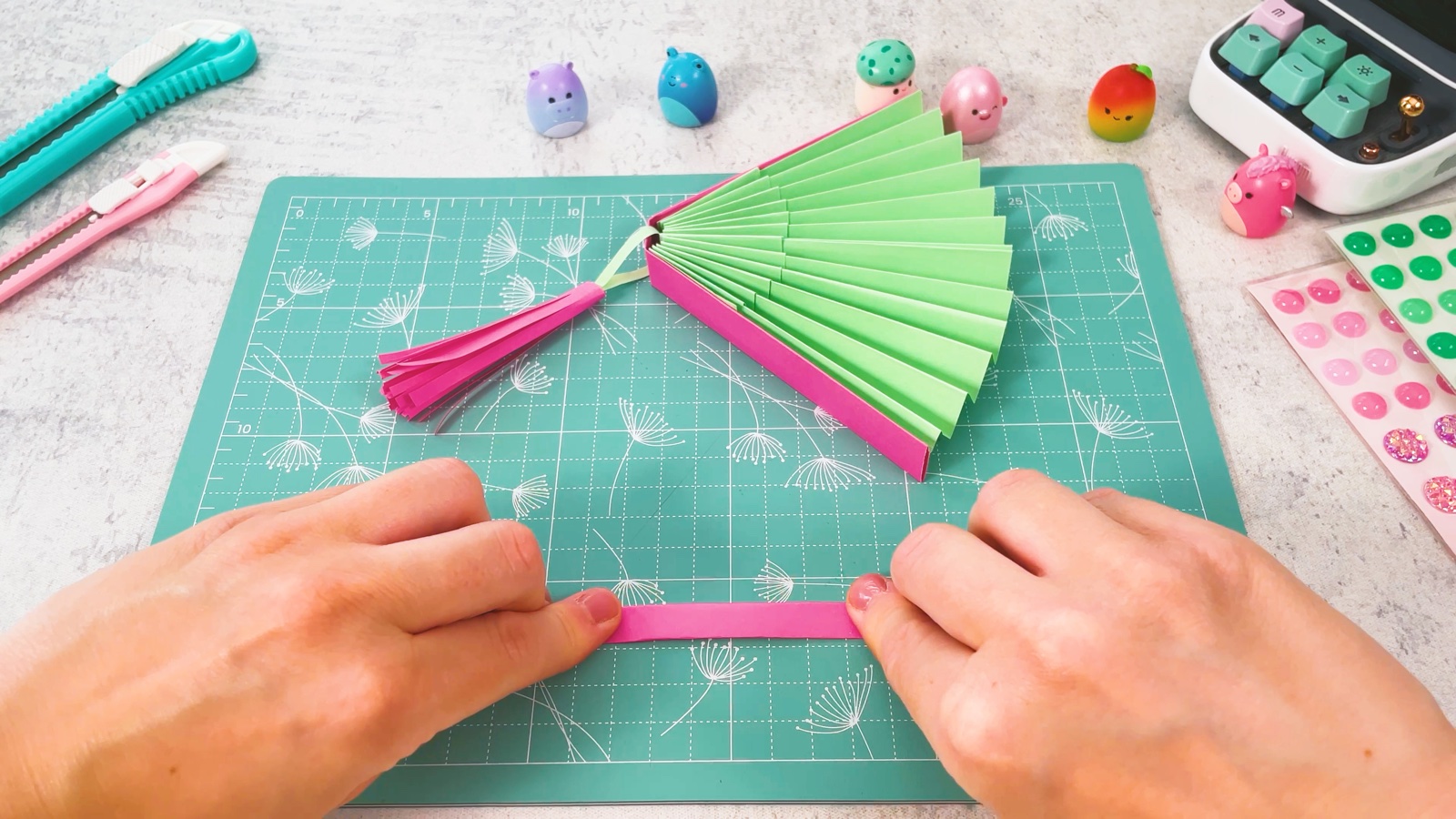 Une petite bande rose étroite pliée sur le tapis de découpe, à côté de l’éventail et de son pompon