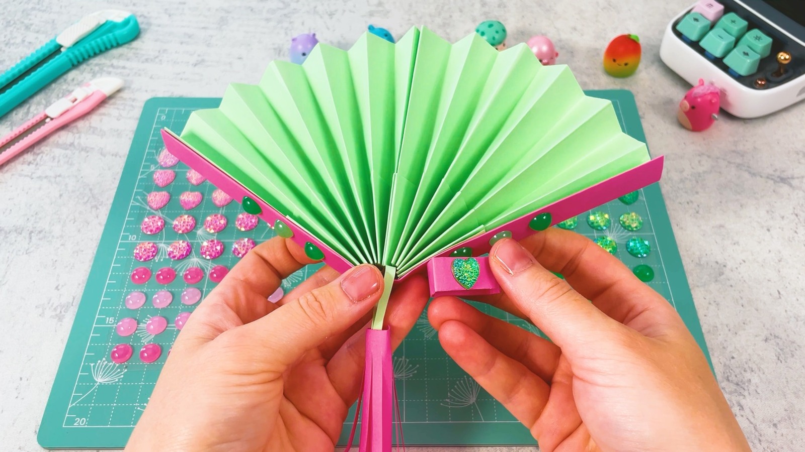 L’éventail en papier terminé, ouvert, avec ses plis verts, ses baguettes roses décorées de strass verts et le pompon rose en dessous, l’anneau brillant tenu dans une main