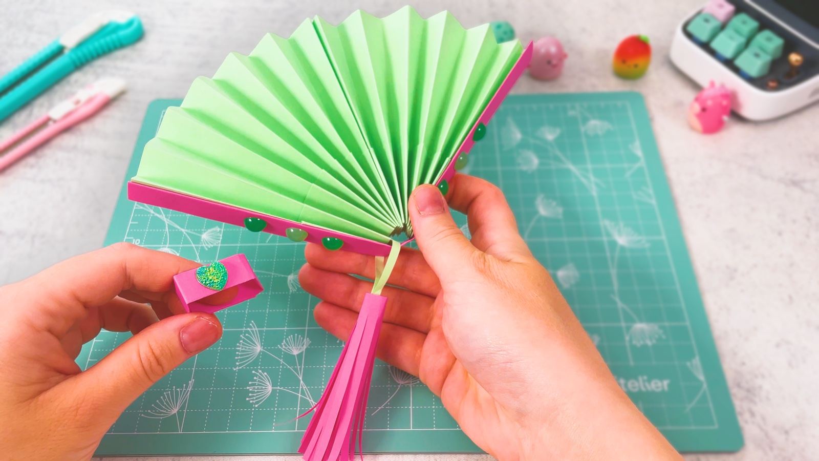 A finished open paper fan with mint green pleats, pink side sticks with gem stickers and a pink paper tassel