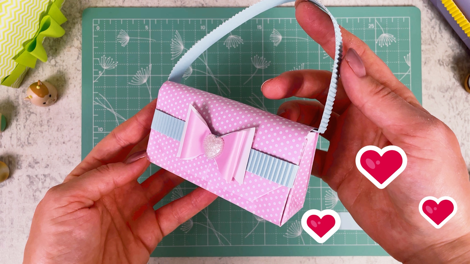 The finished pink polka dot paper purse with a blue handle, a blue band and a pink bow with a heart gem, held in both hands
