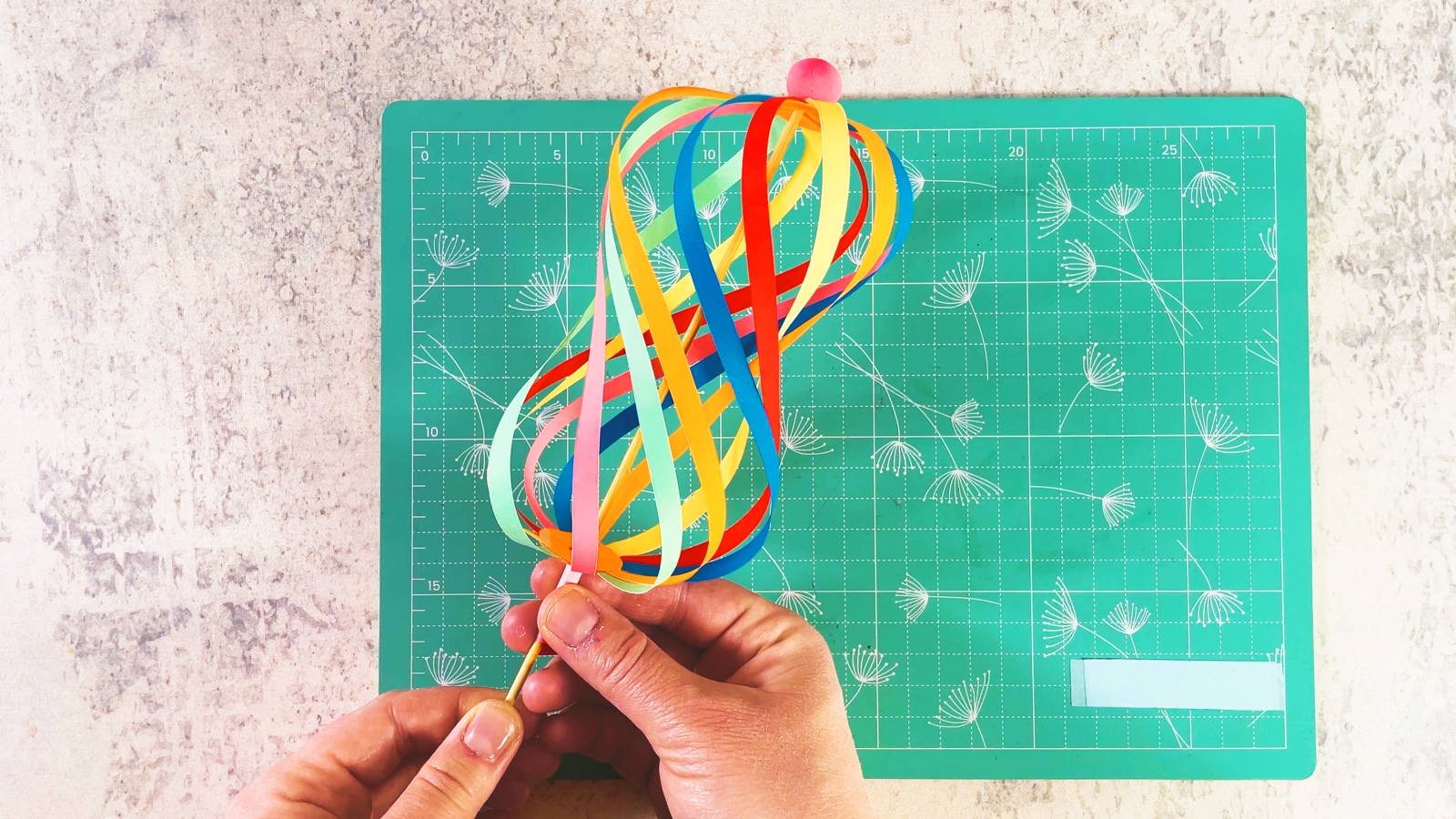 A finished rainbow paper spinner made of twisted paper strips on a stick, held over a mint cutting mat