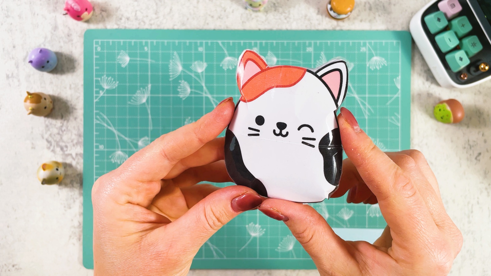 El squishy de gato calicó terminado y esponjado, guiñando un ojo, sostenido sobre el tapete de corte