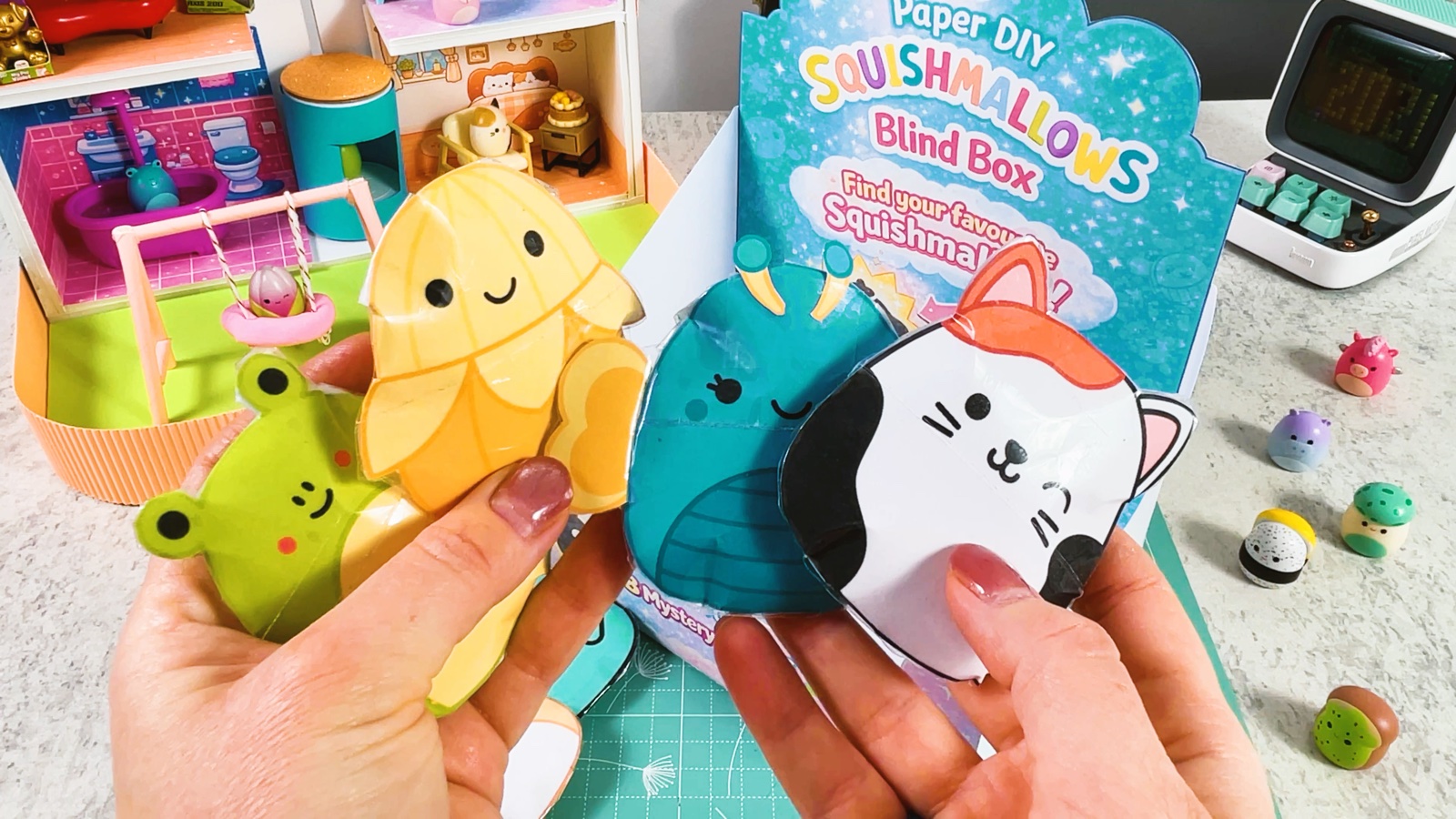 Cuatro Squishmallows de papel terminados, un gato color menta, una mazorca, un gato calicó y una abeja, junto a una bolsa sorpresa