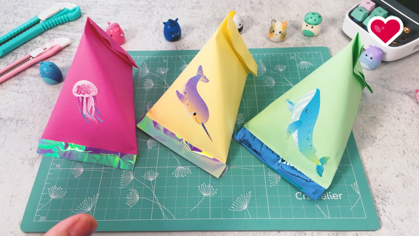 Three finished paper treat bags in pink, yellow and green, with jellyfish, narwhal and whale stickers, standing side by side on a cutting mat