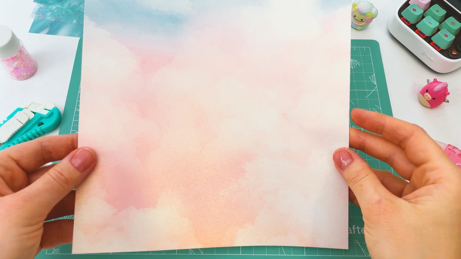 Holding up a sheet of pastel pink and blue cloud paper over a green cutting mat