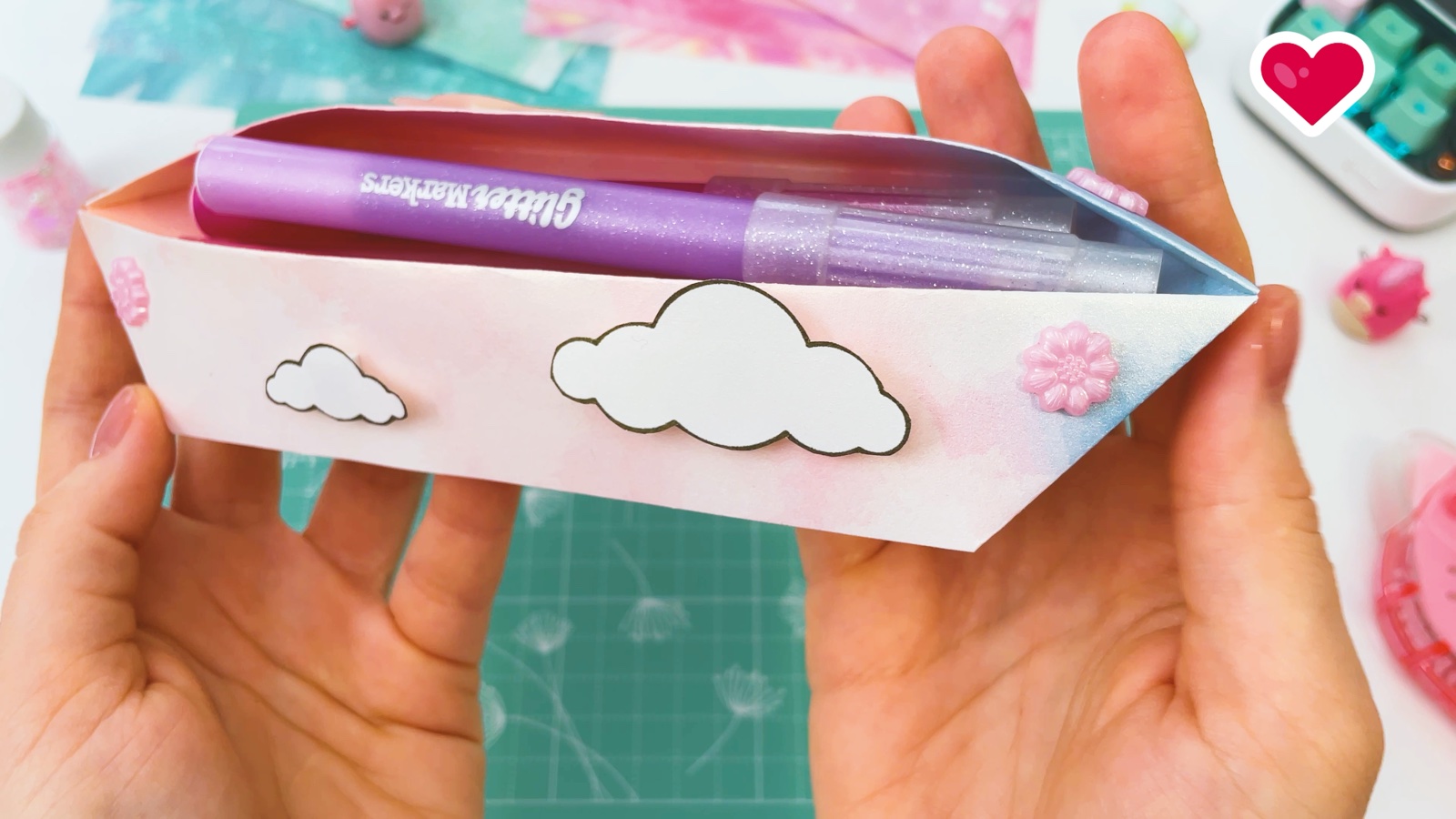 Holding the finished cloudy paper pencil case in both hands, with purple glitter markers inside and pink flower gems at the ends