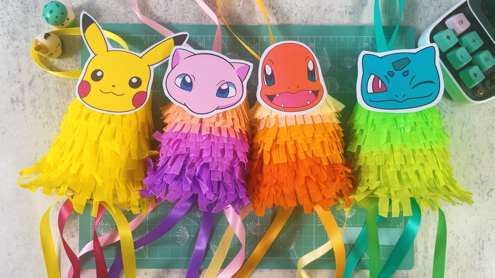 Four finished mini piñatas in a row: yellow Pikachu, purple Mew, orange Charmander and green Bulbasaur, each with long ribbons hanging below