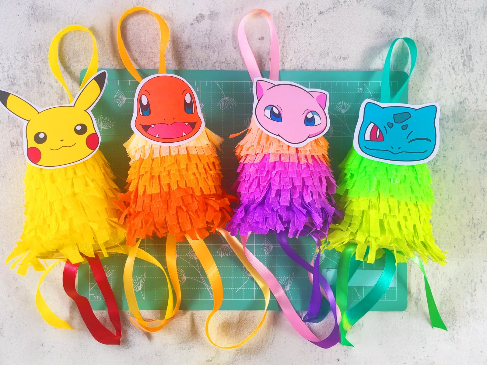 Four finished Pokémon mini piñatas with crepe paper fringe and ribbons, lying on a mint cutting mat