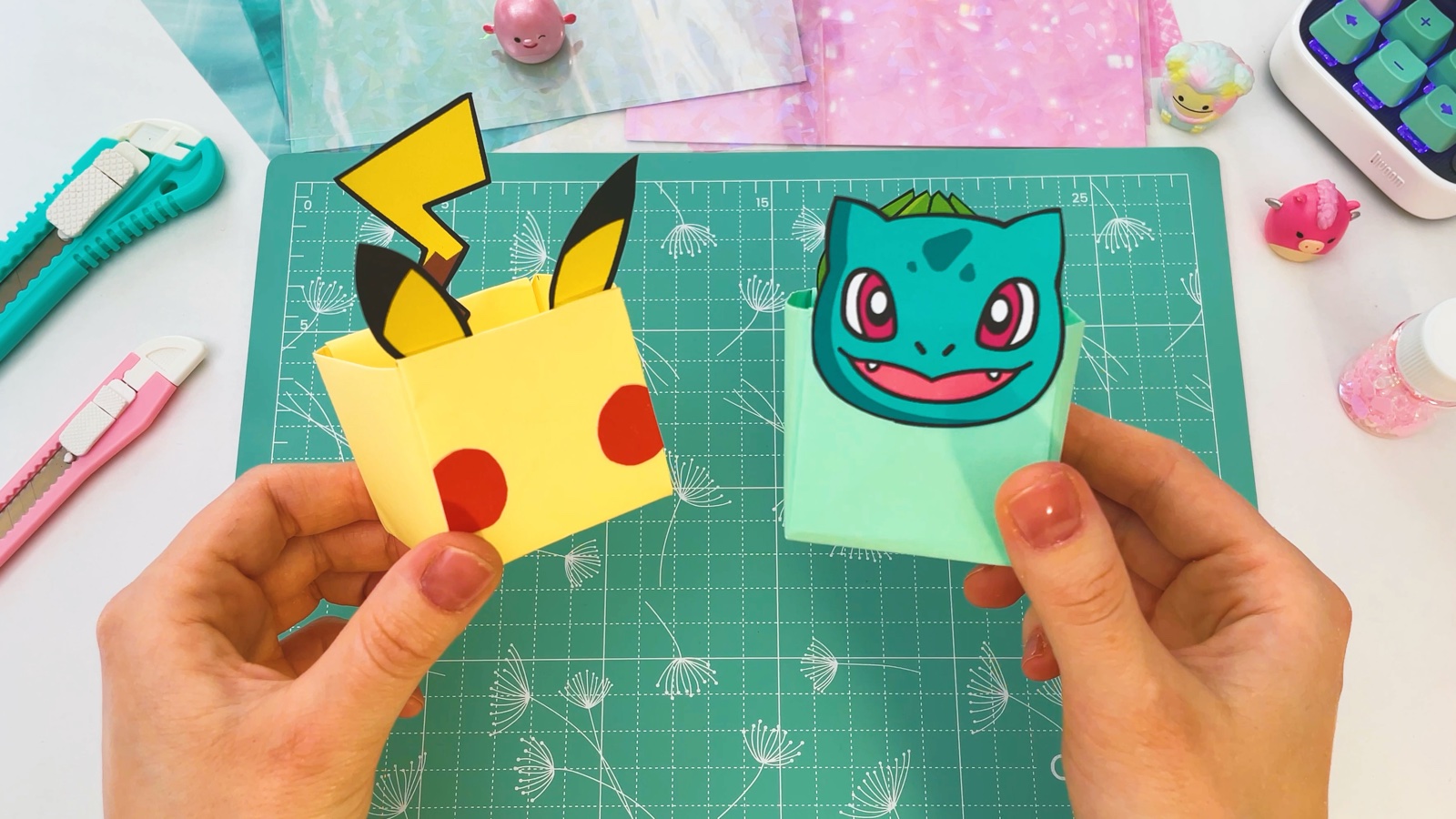 The finished Pikachu box with ears, tail and red cheeks next to the finished Bulbasaur box, held in two hands