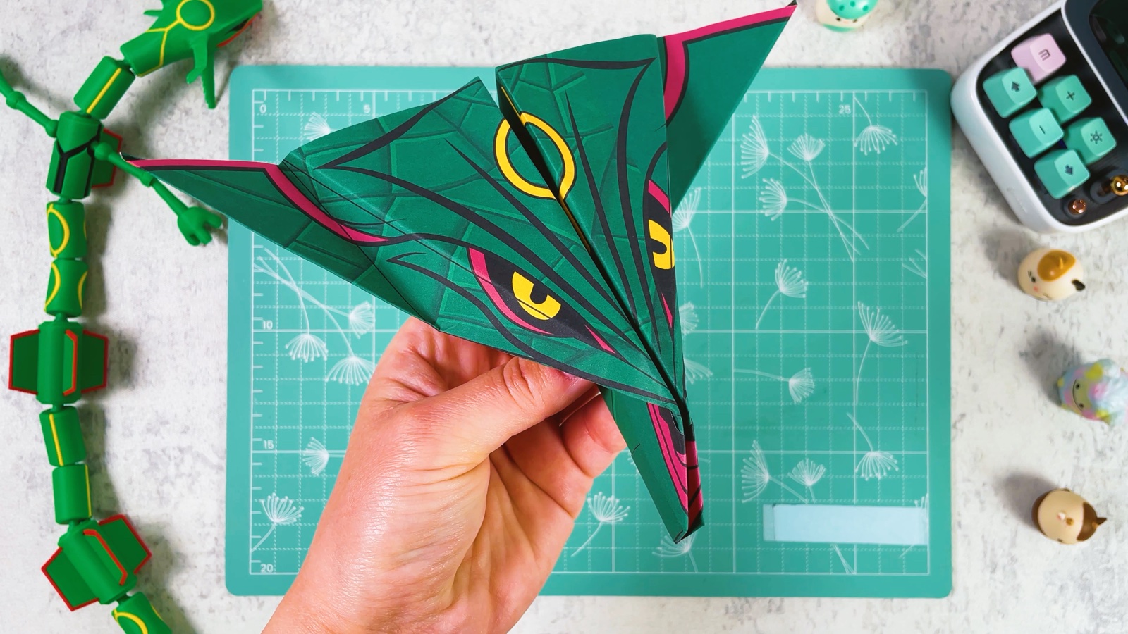 The finished Green Rayquaza paper airplane held from below, with wide pointed wings, yellow rings, red-rimmed eyes and pink edges