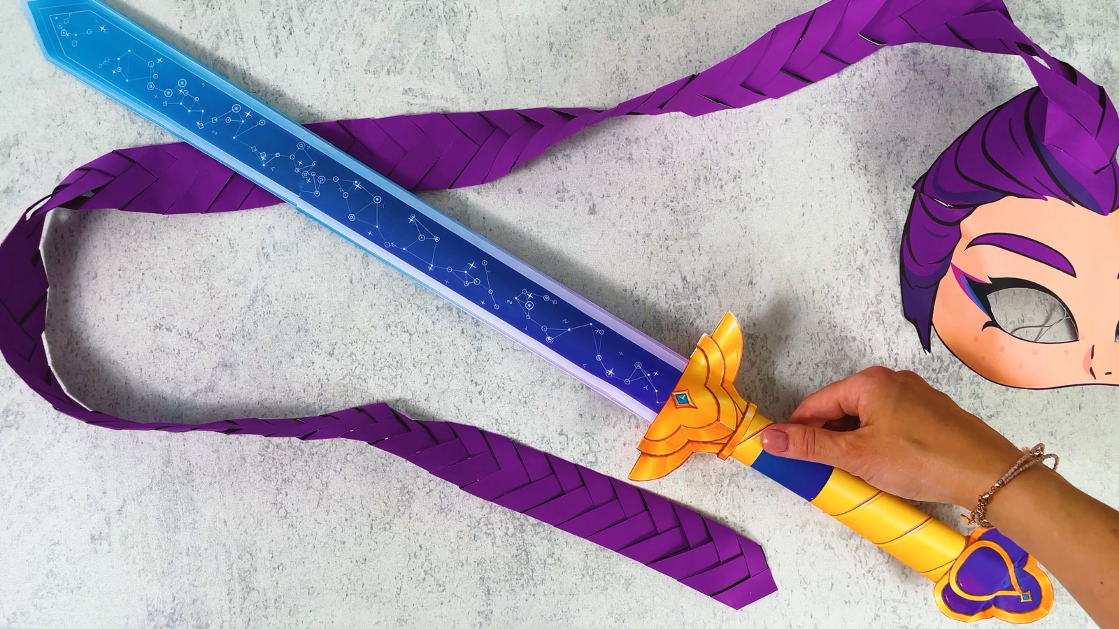 Toute l’épée de Rumi en papier terminée, de la pointe bleue à l’embout violet, posée à côté d’un masque de Rumi en papier avec une longue tresse violette