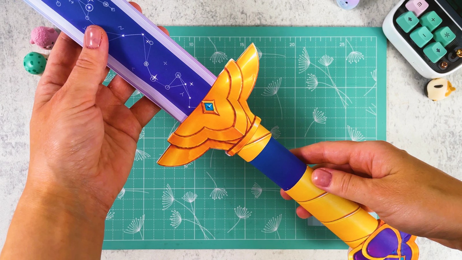 L’épée de Rumi en papier terminée, tenue au-dessus d’un tapis de découpe : la lame violette étoilée, la garde dorée avec une gemme bleue et la poignée dorée et bleue