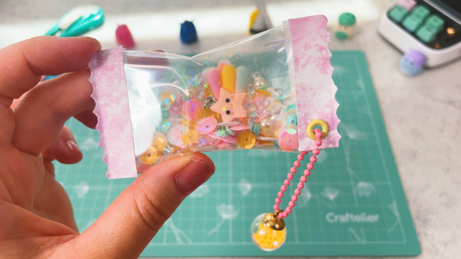 Der fertige Shaker-Anhänger in einer Hand, voller Pastell-Pailletten und mit Sternanhänger, an der Öse hängen die rosa Kugelkette und der gelbe Perlenanhänger
