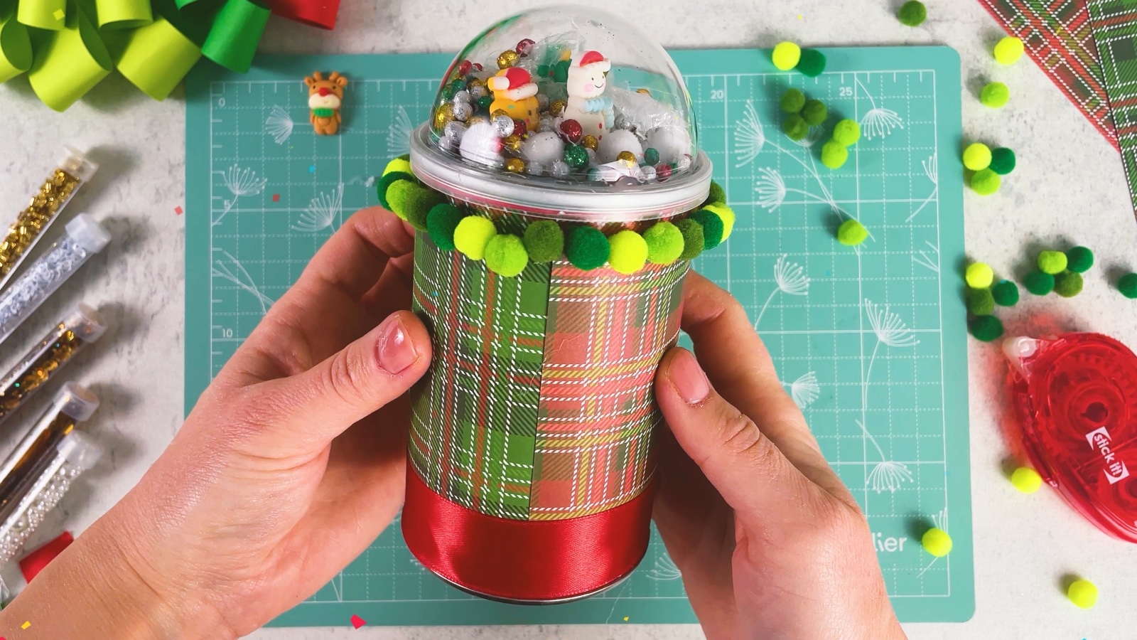 The finished snow globe gift box: plaid can, red ribbon, green pom-pom ring and a clear dome with figurines, beads and snow