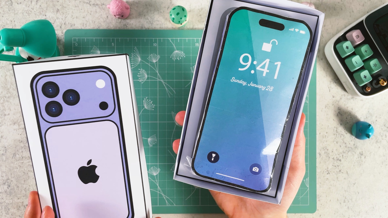 The finished squishy paper iPhone with a blue lock screen, lying in its open white box, next to the box lid with the purple iPhone picture
