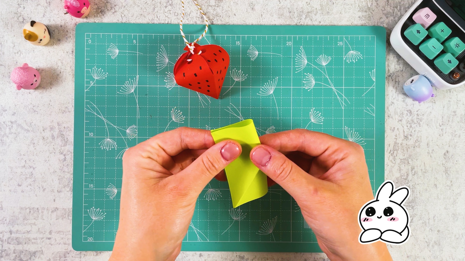 Folding a small green paper square, with the finished red strawberry hanging behind