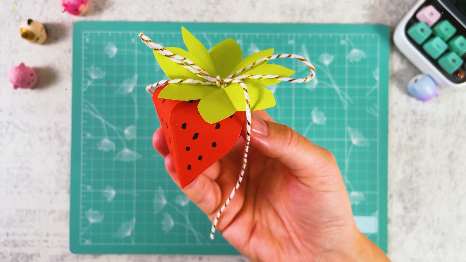 The finished paper strawberry with black seeds, green leaves and a string bow, held in one hand